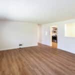 Empty living room with light-colored walls, large window on the left, wood-style flooring, and an open pass-through to a kitchen area with visible appliances and a chandelier in the background.
