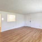 Empty room with light wood flooring, white walls, a white door, and two open interior windows leading to another room with similar flooring. Brightly lit by natural light.
