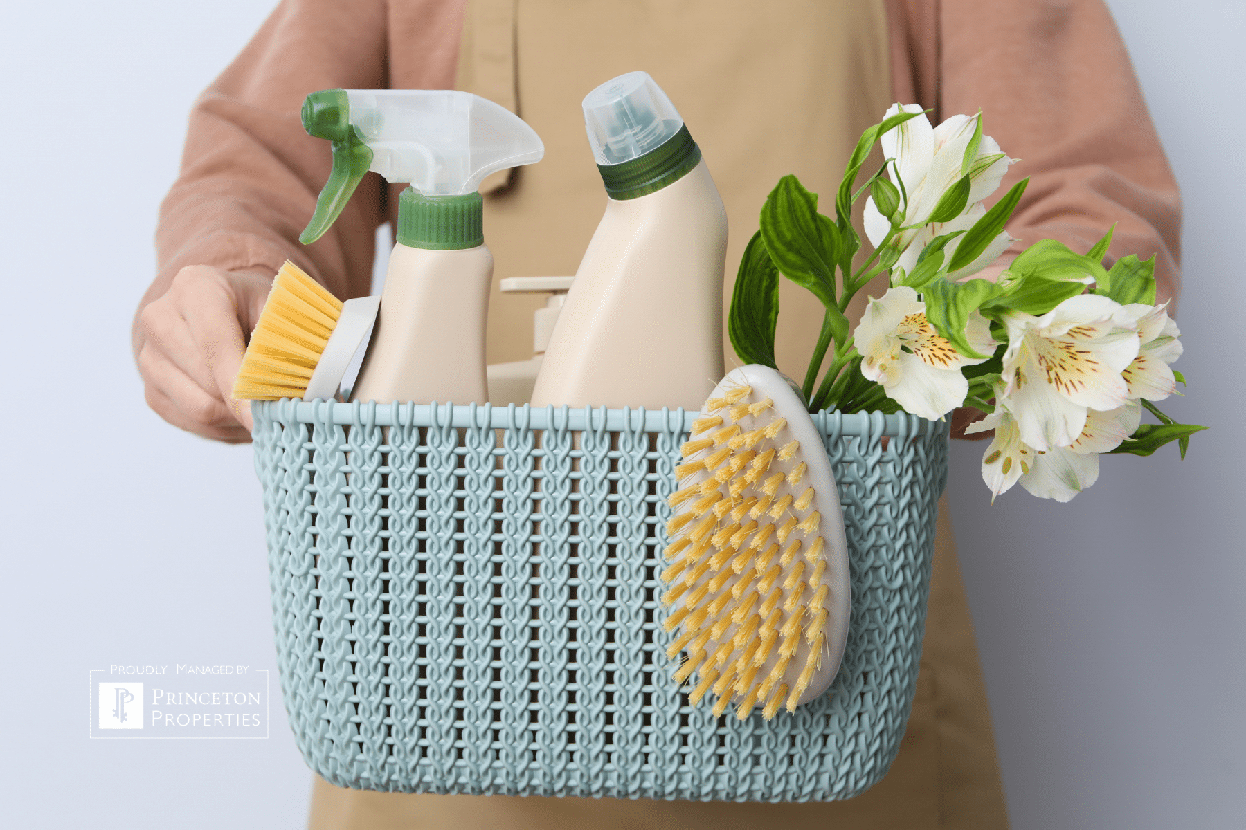 Princeton Properties | A person in a tan apron holds a blue woven basket containing cleaning supplies, a yellow brush, a spray bottle, cleaner bottles, and fresh white flowers. A Princeton Properties logo is visible in the lower left corner. | Spring Cleaning for Renters